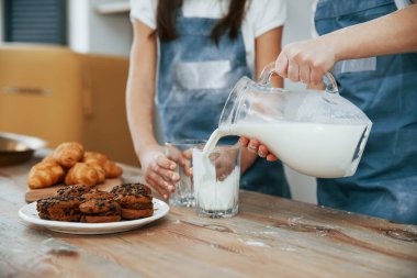Close up view of two little girls in blue chef uniform that pouring milk into glasses on the kitchen with cookies on table.
