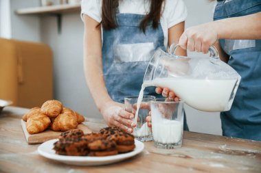 Close up view of two little girls in blue chef uniform that pouring milk into glasses on the kitchen with cookies on table.