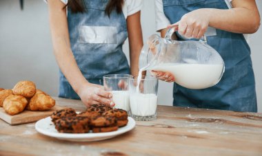 Close up view of two little girls in blue chef uniform that pouring milk into glasses on the kitchen with cookies on table.