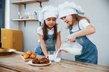 Mavi aşçı üniformalı iki küçük kız mutfaktaki bardaklara süt dolduruyor ve masada kurabiyeler var..