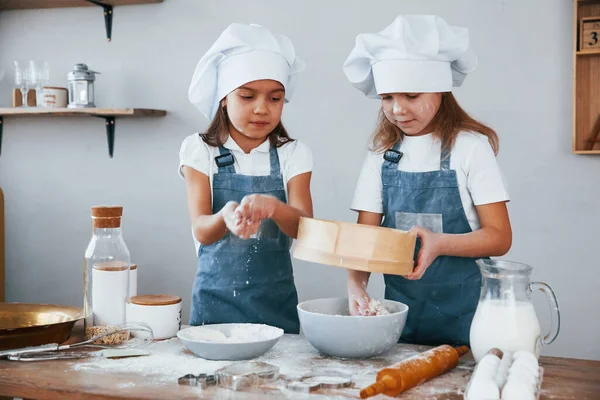 Mavi şef üniformalı iki küçük kız mutfakta elek kullanarak un ile çalışıyorlar..