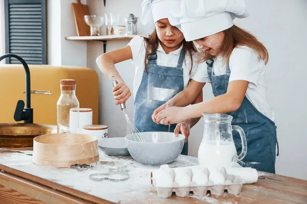 Mavi şef üniformalı iki küçük kız, un ve yumurta kullanarak mutfakta yemek hazırlıyorlar..