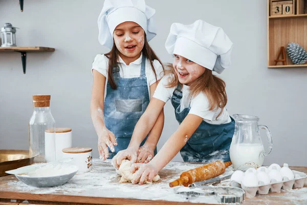 Two little girls in blue chef uniform kneading dough on the kitchen.