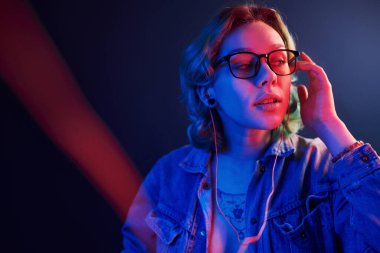 Gözlüklü, yeşil saçlı, kırmızı ve mavi ışıklı genç bir kızın portresi stüdyoda kulaklıkla müzik dinliyor..