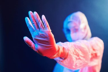 Maskeli hemşire ve beyaz üniformalı neon ışıklı odada duruyor ve dur işareti gösteriyor..