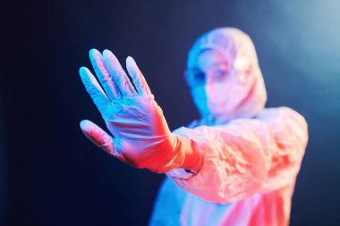 Maskeli hemşire ve beyaz üniformalı neon ışıklı odada duruyor ve dur işareti gösteriyor..