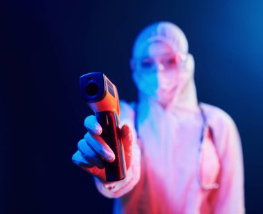 Maskeli hemşire ve beyaz üniformalı adam neon ışıklı odada duruyor ve kızılötesi termometreyi tutuyor. Koronavirüsün yayılmasını durdur..