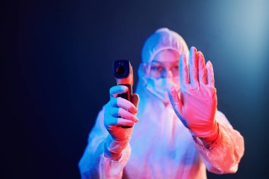 Maskeli hemşire ve beyaz üniformalı adam neon ışıklı odada duruyor ve kızılötesi termometreyi tutuyor. Koronavirüsün yayılmasını durdur..