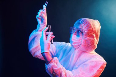 Maskeli hemşire ve beyaz üniformalı neon ışıklı odada duruyor ve elinde numunelerle tüpler tutuyordu..