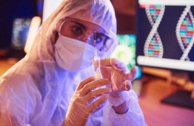 Maskeli hemşire ve beyaz üniformalı, elinde şırınga olan ve neon ışıklı laboratuarda oturan Coronavirus aşısını arayan bilgisayar ve tıbbi ekipmanlarla.