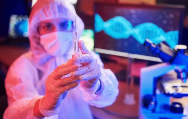 Maskeli hemşire ve beyaz üniformalı, elinde şırınga olan ve neon ışıklı laboratuarda oturan Coronavirus aşısını arayan bilgisayar ve tıbbi ekipmanlarla.