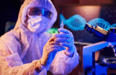 Maskeli hemşire ve beyaz üniformalı, elinde şırınga olan ve neon ışıklı laboratuarda oturan Coronavirus aşısını arayan bilgisayar ve tıbbi ekipmanlarla.