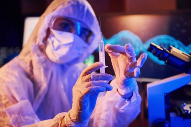 Maskeli hemşire ve beyaz üniformalı, elinde şırınga olan ve neon ışıklı laboratuarda oturan Coronavirus aşısını arayan bilgisayar ve tıbbi ekipmanlarla.