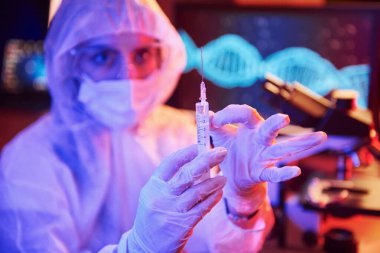 Maskeli hemşire ve beyaz üniformalı, elinde şırınga olan ve neon ışıklı laboratuarda oturan Coronavirus aşısını arayan bilgisayar ve tıbbi ekipmanlarla.