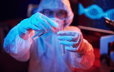 Maskeli hemşire ve beyaz üniformalı, elinde sıvı tüple neon ışıklı laboratuarda oturmuş Coronavirus aşısını arıyor..