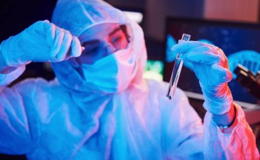 Maskeli hemşire ve beyaz üniformalı, elinde sıvı tüple neon ışıklı laboratuarda oturmuş Coronavirus aşısını arıyor..