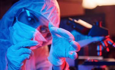 Maskeli hemşire ve beyaz üniformalı, elinde sıvı tüple neon ışıklı laboratuarda oturmuş Coronavirus aşısını arıyor..