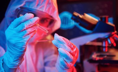 Maskeli hemşire ve beyaz üniformalı, elinde sıvı tüple neon ışıklı laboratuarda oturmuş Coronavirus aşısını arıyor..