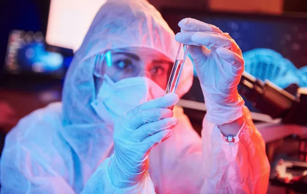 Maskeli hemşire ve beyaz üniformalı, elinde sıvı tüple neon ışıklı laboratuarda oturmuş Coronavirus aşısını arıyor..