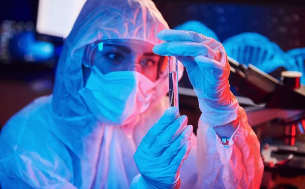 Maskeli hemşire ve beyaz üniformalı, elinde sıvı tüple neon ışıklı laboratuarda oturmuş Coronavirus aşısını arıyor..