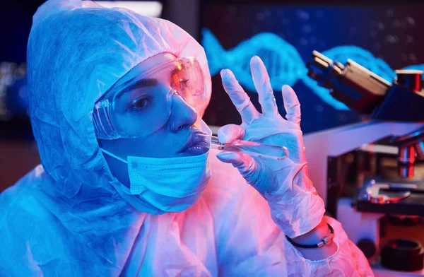 Maskeli hemşire ve beyaz üniformalı, elinde sıvı tüple neon ışıklı laboratuarda oturmuş Coronavirus aşısını arıyor..