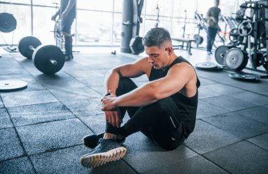 Spor elbiseli güçlü genç adam spor salonunda spor yapıyor, oturuyor ve mola veriyor..