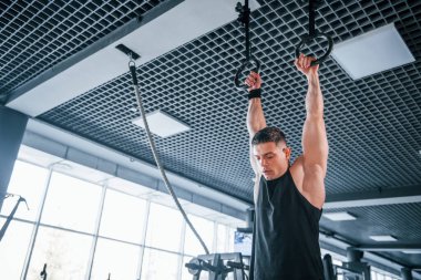 Spor kıyafetleri içindeki güçlü genç adam spor salonunda jimnastik yüzükleri üzerinde egzersiz yapıyor..