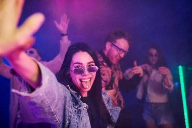 Gece kulübünde renkli lazer ışıklarıyla eğlenen gençlerin önünde elinde uyuşturucu hapları tutan genç bir kız..