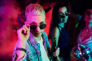 Güneş gözlüklü hippi adam ve bir şişe alkolle gece kulübünde renkli lazer ışıklarıyla eğlenen gençlerin önünde kameraya poz veriyor..