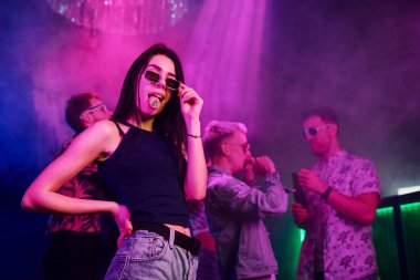 Gece kulübünde renkli lazer ışıklarıyla eğlenen gençlerin önünde elinde uyuşturucu hapları tutan genç bir kız..