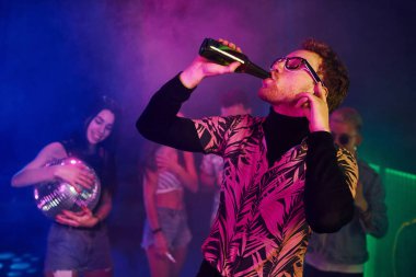 Gözlüklü adam bira içiyor. Gençler gece kulübünde renkli lazer ışıklarıyla eğleniyorlar..