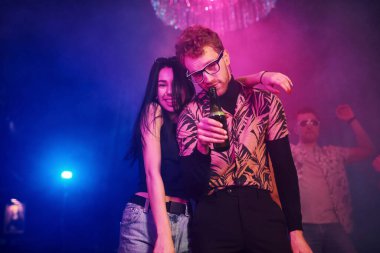 Gözlüklü adam bira içiyor. Gençler gece kulübünde renkli lazer ışıklarıyla eğleniyorlar..