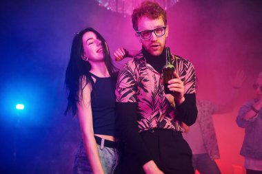 Gözlüklü adam bira içiyor. Gençler gece kulübünde renkli lazer ışıklarıyla eğleniyorlar..