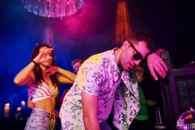 Gece kulübünde renkli lazer ışıklarıyla eğlenen genç insanların önünde çitlere yaslanan sarhoş adam..