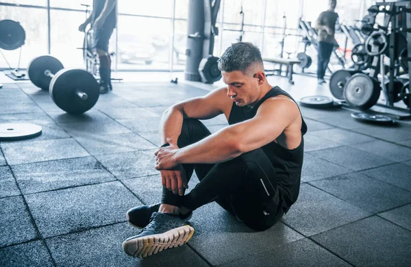 Spor elbiseli güçlü genç adam spor salonunda spor yapıyor, oturuyor ve mola veriyor..
