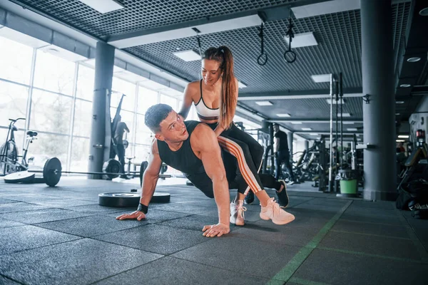 Kadın şınav çeken adamın üstüne oturur. Spor salonunda birlikte egzersiz yapan genç insanlar..