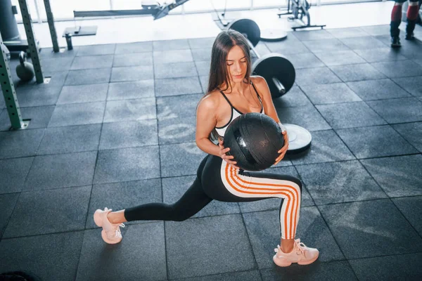 Spor elbiseli genç bir kadın spor salonunda çömeliyor ve topu elinde tutuyor..