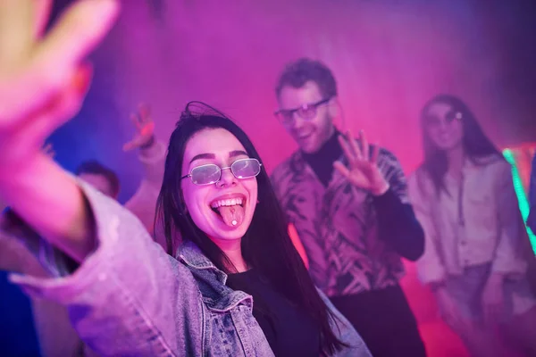 Young girl holding drug pills on togue in front of young people that having fun in night club with colorful laser lights.