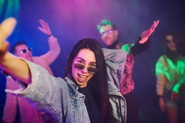 Young girl holding drug pills on togue in front of young people that having fun in night club with colorful laser lights.