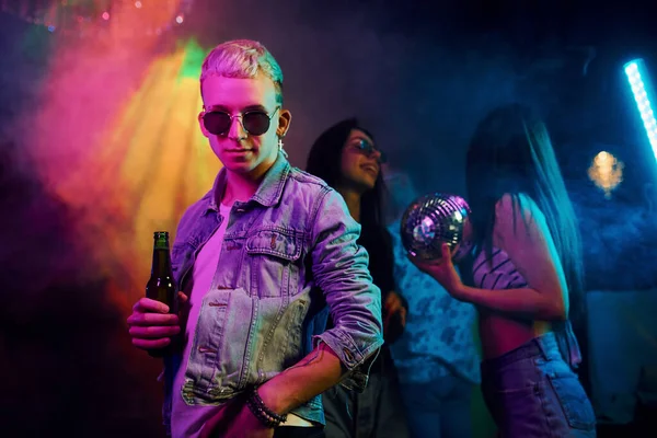 Güneş gözlüklü hippi adam ve bir şişe alkolle gece kulübünde renkli lazer ışıklarıyla eğlenen gençlerin önünde kameraya poz veriyor..