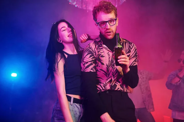Gözlüklü adam bira içiyor. Gençler gece kulübünde renkli lazer ışıklarıyla eğleniyorlar..