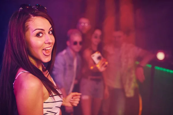 Young girl holding drug pills on togue in front of young people that having fun in night club with colorful laser lights.