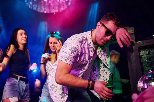 Gece kulübünde renkli lazer ışıklarıyla eğlenen genç insanların önünde çitlere yaslanan sarhoş adam..