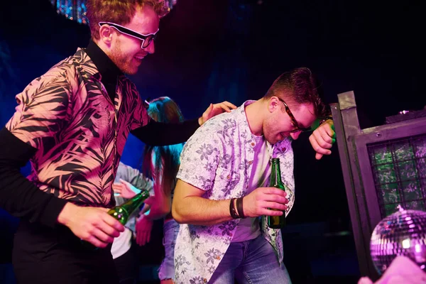 Gece kulübünde renkli lazer ışıklarıyla eğlenen genç insanların önünde çitlere yaslanan sarhoş adam..