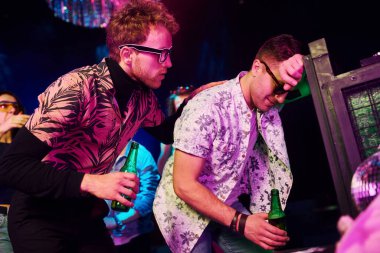 Gece kulübünde renkli lazer ışıklarıyla eğlenen genç insanların önünde çitlere yaslanan sarhoş adam..