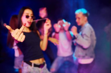 Gözlüklü kadın orta parmak gösteriyor. Gençler gece kulübünde renkli lazer ışıklarıyla eğleniyorlar..