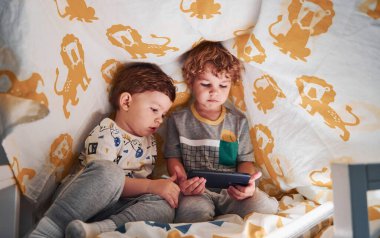 İki küçük çocuk dinleniyor ve akıllı telefonlarıyla birlikte battaniyenin altında eğleniyorlar..