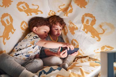 İki küçük çocuk dinleniyor ve akıllı telefonlarıyla birlikte battaniyenin altında eğleniyorlar..