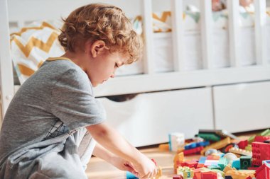 Cute little boy sitting and have fun indoors in the bedroom with plastic construction set.