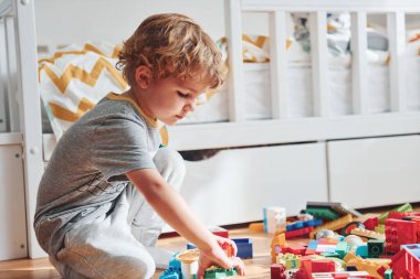 Cute little boy sitting and have fun indoors in the bedroom with plastic construction set.